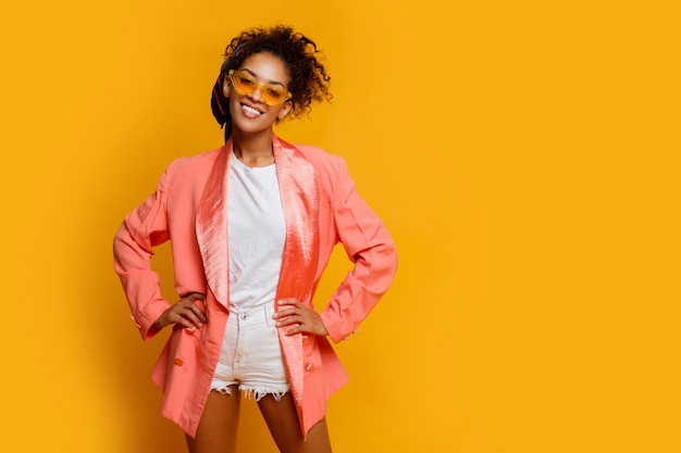 Confiante femme noire souriante dans une élégante veste rose posant à l'intérieur sur fond jaune.