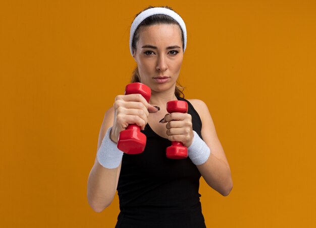 Confiante jeune femme assez sportive portant un bandeau et des bracelets tenant des haltères faisant un geste de boxe