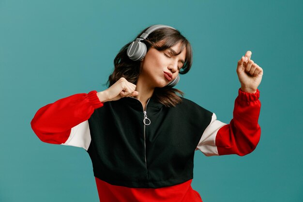 Confiante jeune femme caucasienne portant des écouteurs gardant les poings dans l'air dansant avec les yeux fermés isolés sur fond bleu
