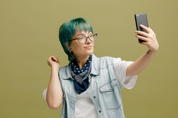 Confiante jeune femme portant des lunettes bandana sur le cou en gardant la main dans l'air étirant le téléphone portable en prenant selfie isolé sur fond vert olive