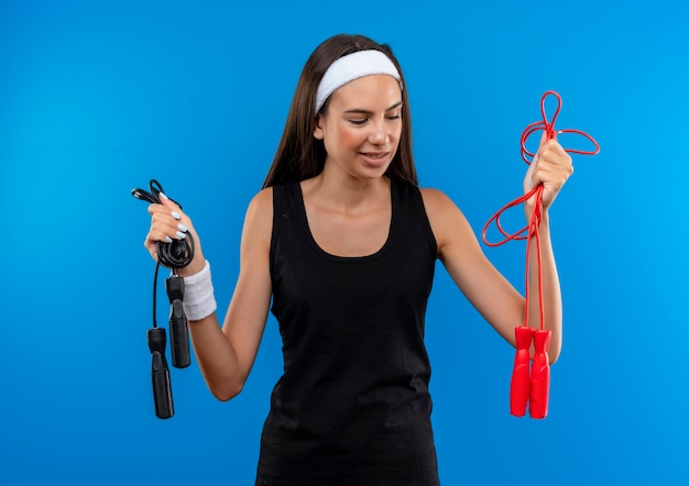 Confiante jeune fille assez sportive portant un bandeau et un bracelet tenant et regardant des cordes à sauter sur un mur bleu