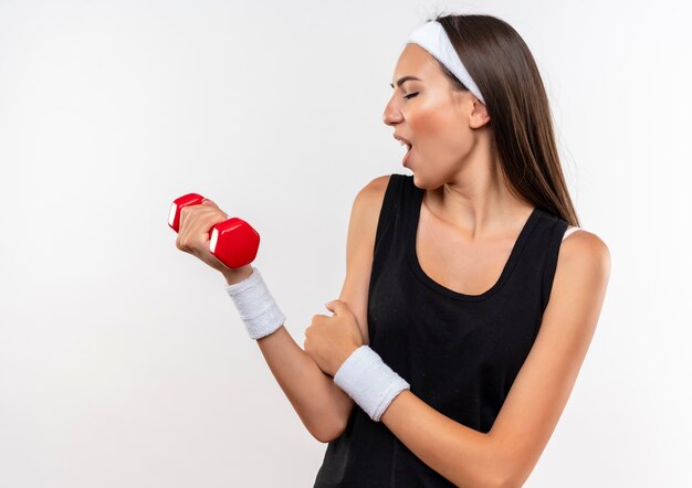 Confiante jeune fille assez sportive portant un bandeau et un bracelet tenant et regardant l'haltère et mettant la main sur le bras isolé sur un mur blanc