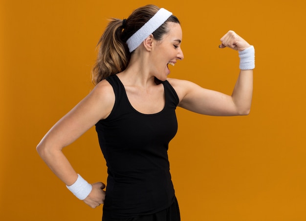 Confiante jeune fille assez sportive portant un bandeau et des bracelets faisant un geste fort en regardant ses muscles isolés sur un mur orange