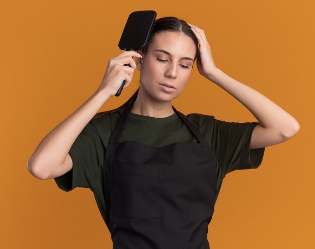 Confiante jeune fille de barbier brune en uniforme peignant les cheveux et met la main sur la tête isolée sur un mur orange avec espace de copie