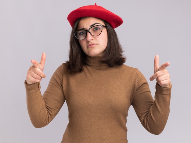 Confiante jeune jolie fille caucasienne avec chapeau de béret et lunettes optiques pointant vers la caméra à deux mains