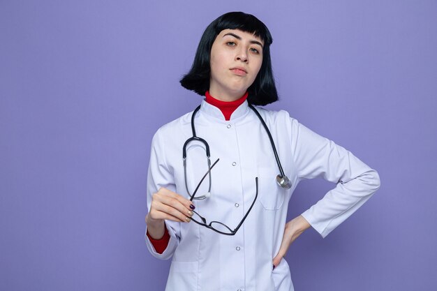 Confiante jeune jolie fille caucasienne en uniforme de médecin avec stéthoscope tenant des lunettes optiques et