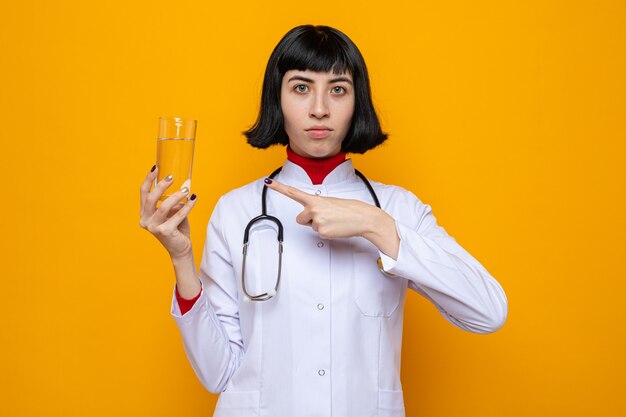Confiante jeune jolie fille caucasienne en uniforme de médecin avec stéthoscope tenant un verre d'eau