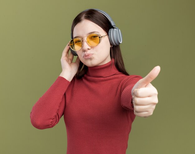 Confiante jeune jolie fille portant des lunettes de soleil et des écouteurs touchant des écouteurs montrant le pouce vers le haut isolé sur un mur vert olive