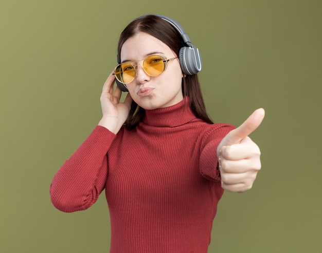 Confiante jeune jolie fille portant des lunettes de soleil et des écouteurs touchant des écouteurs montrant le pouce vers le haut isolé sur un mur vert olive