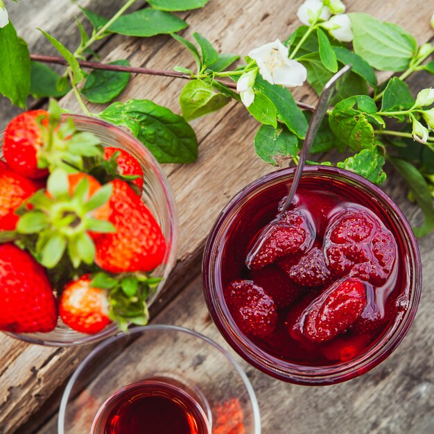 Confiture de fraises avec cuillère, thé en verre, fraises, branche de fleur dans une assiette sur table en bois, mise à plat.