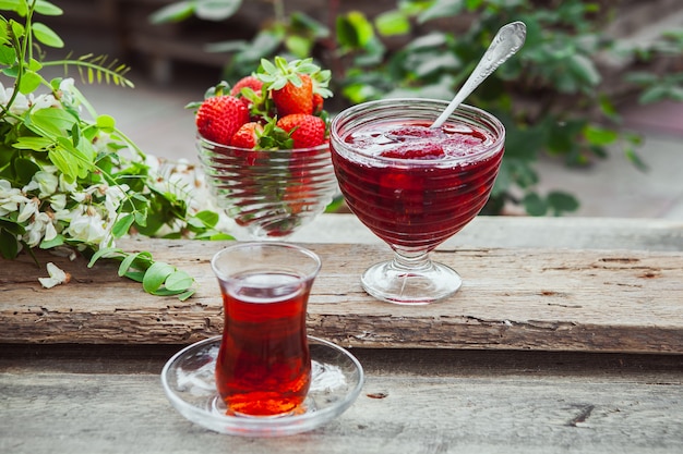 Confiture de fraises dans une assiette avec cuillère, un verre de thé, fraises, vue de dessus des plantes sur table en bois et trottoir