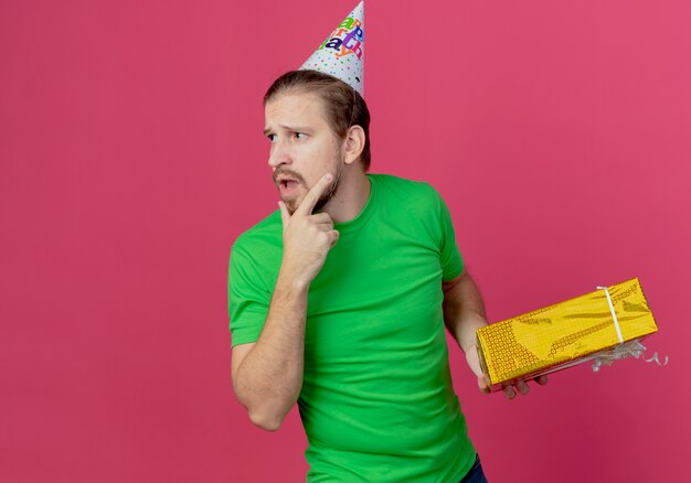 Confus bel homme en chapeau d'anniversaire tient une boîte-cadeau à côté isolé sur mur rose