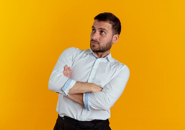 Confus bel homme se tient avec les bras croisés à côté isolé sur mur orange