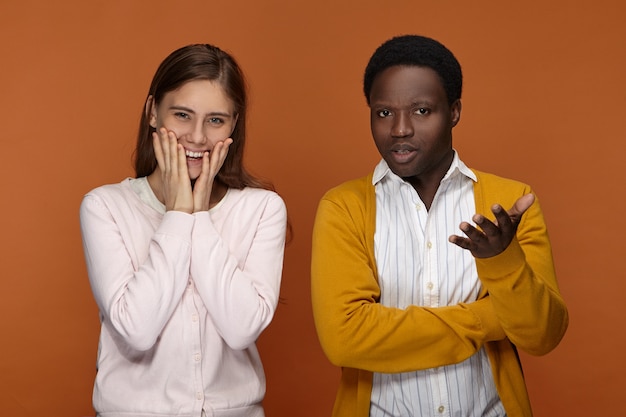 confus désemparé jeune homme afro-américain faisant des gestes, étant à perte tandis que belle femme caucasienne positive tenant les mains sur ses joues et souriant joyeusement, se sentir excité