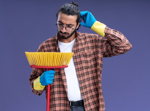 Confus jeune beau mec de nettoyage portant un t-shirt et des gants tenant une vadrouille se gratter la tête isolée sur le mur bleu