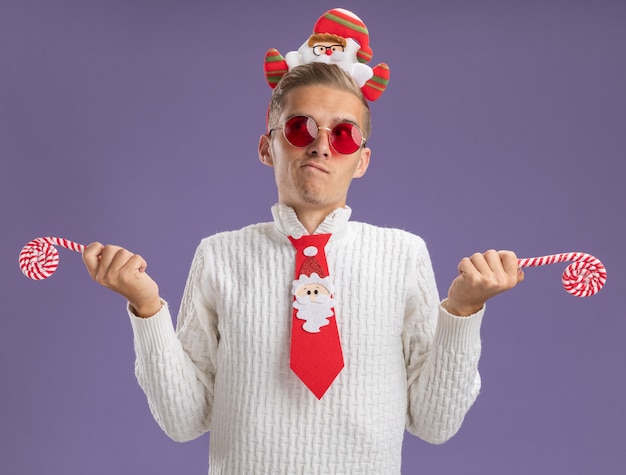 Photo gratuite confus jeune beau mec portant bandeau de père noël et cravate avec des lunettes tenant des cannes de noël à côté isolé sur fond violet