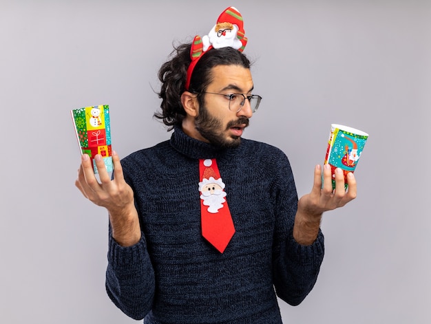 Photo gratuite confus jeune beau mec portant une cravate de noël avec cerceau de cheveux tenant et regardant des tasses de noël isolé sur mur blanc