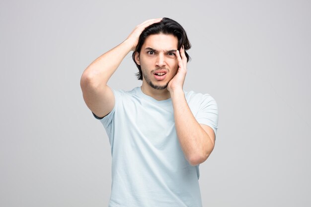 confus jeune bel homme regardant la caméra en gardant les mains sur la tête isolé sur fond blanc