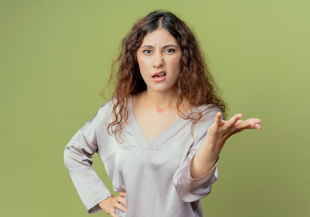 Confus jeune employé de bureau jolie femme tenant la main et mettant la main sur la hanche isolé sur mur vert olive