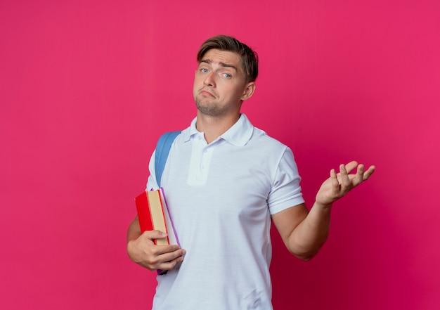 Confus jeune étudiant beau mâle portant un sac à dos tenant des livres et répandre la main isolée sur un mur rose