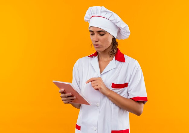 Confus jeune femme cuisinier portant l'uniforme de chef tenant et regardant le cahier sur un mur jaune isolé avec copie espace