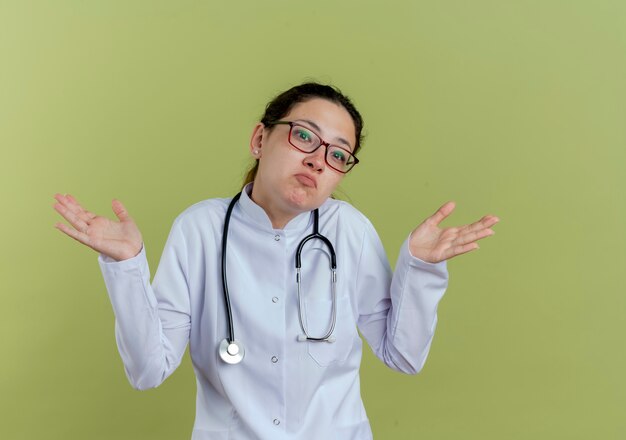 Confus jeune femme médecin portant une robe médicale et un stéthoscope avec des lunettes écartant les mains isolées