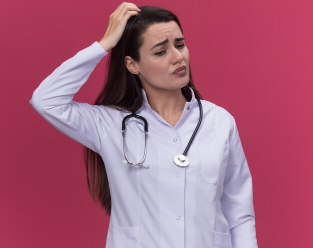 Confus jeune femme médecin portant une robe médicale avec stéthoscope met la main sur la tête et regarde le rose