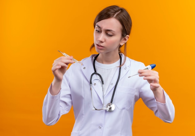 Confus jeune femme médecin portant une robe médicale et un stéthoscope et tenant des thermomètres en les regardant sur l'espace orange isolé