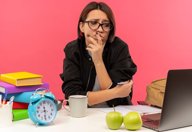 Confus jeune fille étudiante portant des lunettes assis au bureau en regardant le côté mettant la main sur le menton et les lèvres et un autre sur le bras avec un stylo à faire ses devoirs isolés sur rose