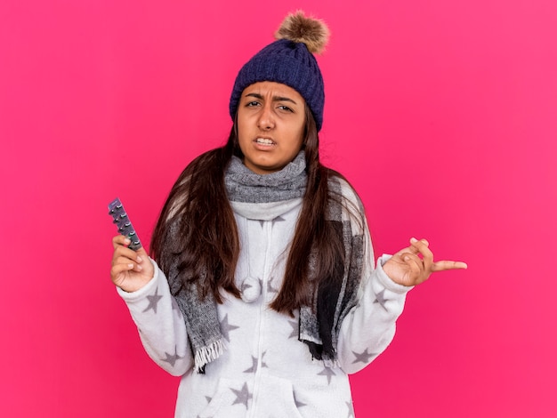 Confus jeune fille malade portant chapeau d'hiver avec foulard tenant des pilules et des points sur le côté isolé sur fond rose avec espace de copie