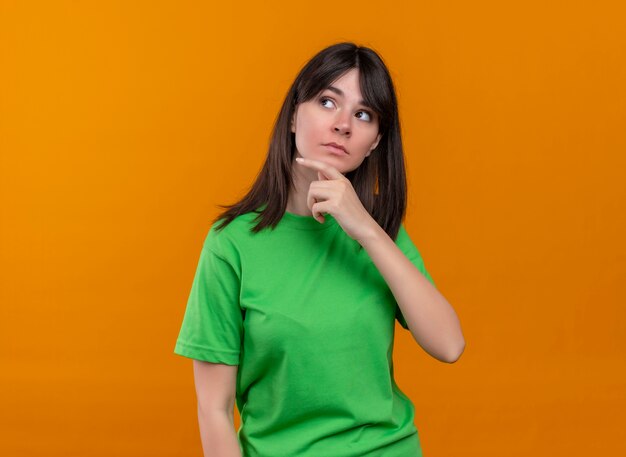 Confus jeune fille de race blanche en chemise verte met la main sur le menton et regarde sur le côté sur fond orange isolé