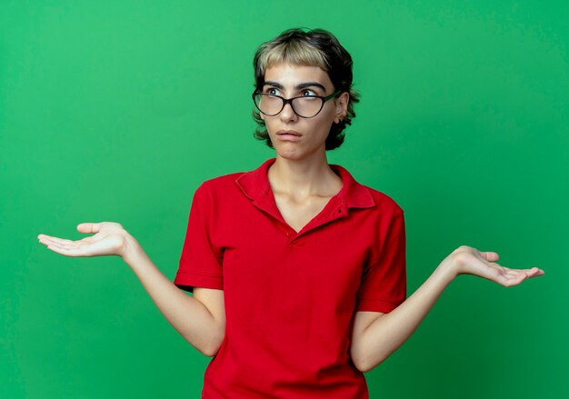 Confus jeune fille de race blanche avec coupe de cheveux de lutin portant des lunettes et montrant les mains vides à côté isolé sur fond vert