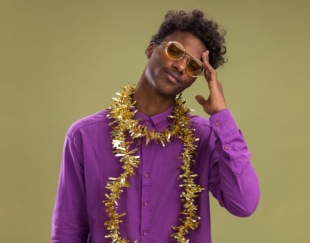 Confus jeune homme afro-américain portant des lunettes avec guirlande de guirlandes autour du cou regardant la caméra toucher la tête isolé sur fond vert olive