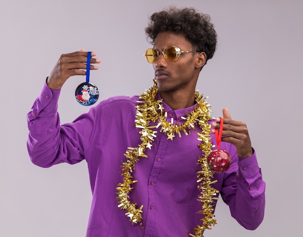 Confus jeune homme afro-américain portant des lunettes avec guirlande de guirlandes autour du cou tenant des boules de Noël en regardant l'un d'eux isolé sur fond blanc