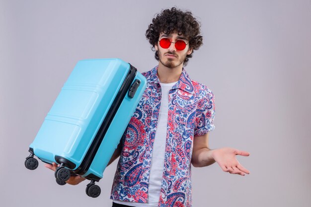 Confus jeune homme beau voyageur bouclé portant des lunettes de soleil tenant valise et montrant la main vide sur un espace blanc isolé avec copie espace