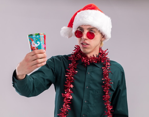 Confus jeune homme blond portant Bonnet de Noel et lunettes avec guirlande de guirlandes autour du cou qui s'étend de la coupe de Noël en plastique en le regardant isolé sur fond blanc