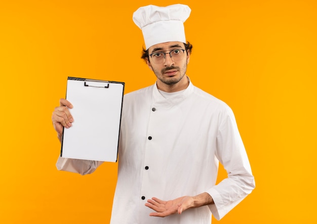 Confus jeune homme cuisinier portant l'uniforme de chef et des lunettes tenant le presse-papiers