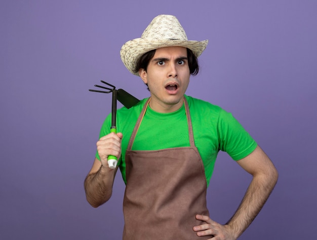 Confus jeune homme jardinier en uniforme portant chapeau de jardinage tenant un râteau houe et mettant la main sur la hanche