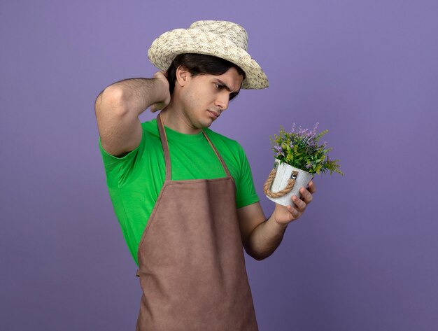 Confus jeune homme jardinier en uniforme portant chapeau de jardinage tenant et regardant la fleur en pot de fleurs en mettant la main sur le cou isolé sur violet