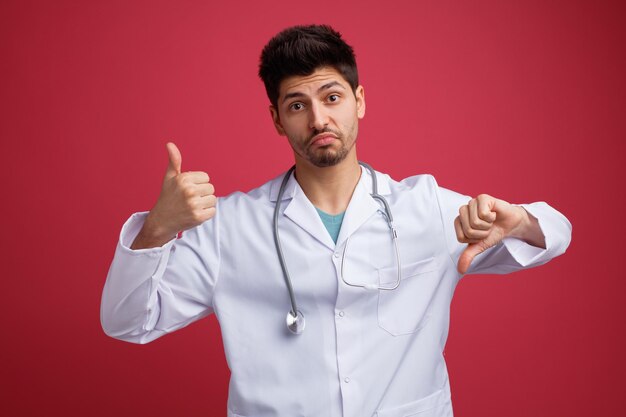 Confus jeune homme médecin portant un uniforme médical et un stéthoscope autour du cou regardant la caméra montrant les pouces de haut en bas isolé sur fond rouge