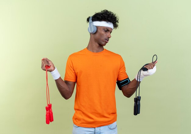 Confus jeune homme sportif afro-américain portant bandeau et bracelet et brassard de téléphone avec des écouteurs tenant et regardant la corde à sauter isolé sur fond vert