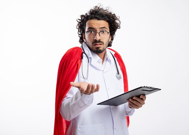 Confus jeune homme de super-héros caucasien à lunettes optiques portant l'uniforme du médecin avec manteau rouge et avec stéthoscope autour du cou tient le presse-papiers et les points avec la main