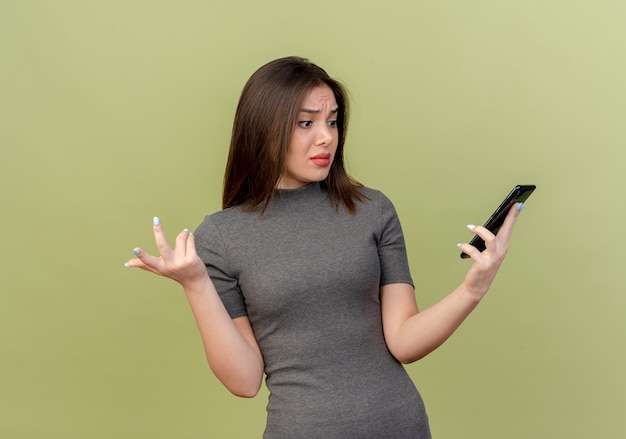 Confus jeune jolie femme tenant et regardant le téléphone mobile et montrant la main vide isolée sur fond vert olive