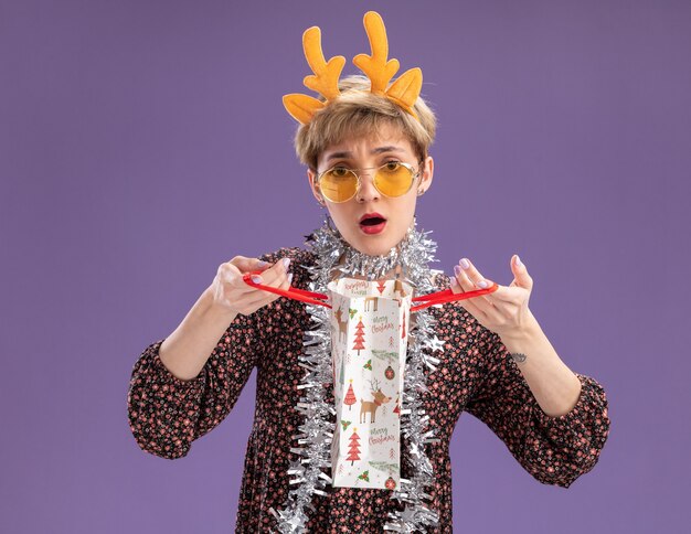 Confus jeune jolie fille portant bandeau de bois de renne et guirlande de guirlandes autour du cou avec des lunettes tenant le sac de cadeau de Noël regardant la caméra isolée sur fond violet