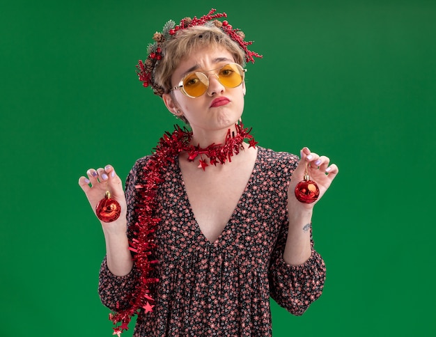 Photo gratuite confus jeune jolie fille portant couronne de tête de noël et guirlande de guirlandes autour du cou avec des lunettes tenant des boules de noël isolé sur mur vert