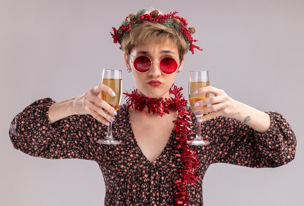 Confus jeune jolie fille portant couronne de tête de Noël et guirlande de guirlandes autour du cou avec des lunettes tenant deux verres de champagne regardant la caméra isolée sur fond blanc