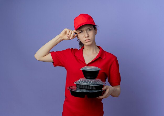 Confus jeune jolie livreuse portant un uniforme rouge et une casquette tenant des contenants de nourriture touchant l'oeil isolé sur fond violet avec espace de copie