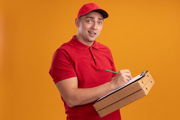 Confus jeune livreur en uniforme avec capuchon tenant des boîtes de pizza et écrire quelque chose sur le presse-papiers isolé sur mur orange