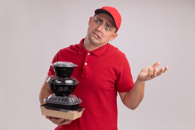 Confus jeune livreur vêtu d'un uniforme avec capuchon tenant des contenants de nourriture propagation main isolé sur mur blanc