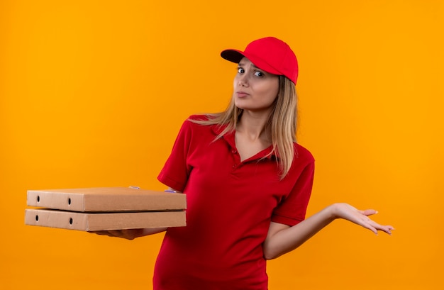 Confus jeune livreuse portant l'uniforme rouge et cap tenant la boîte à pizza et répandre la main isolée sur le mur orange